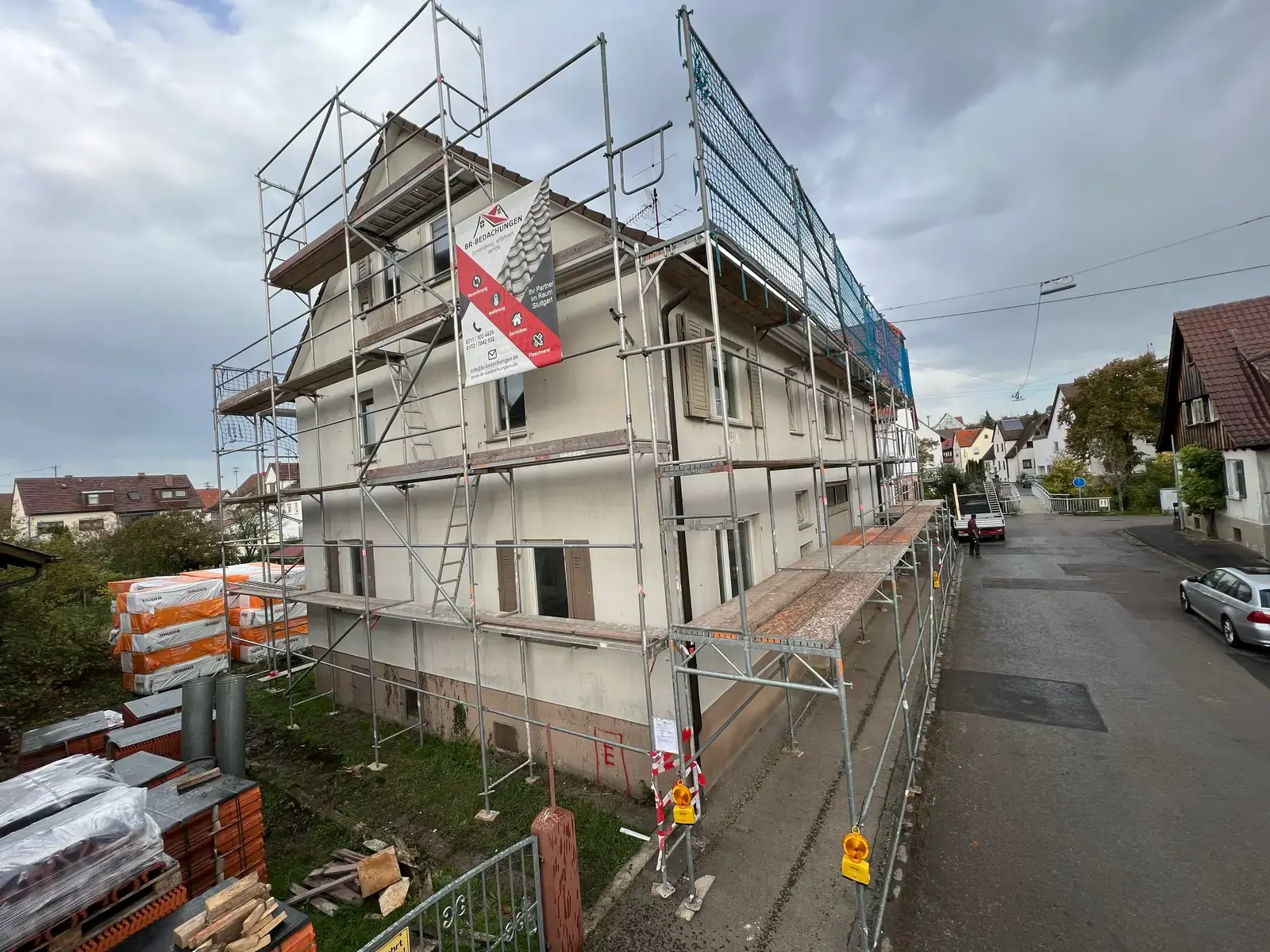 Fassadengerüst von BR-Bedachungen mit Firmenbanner und Fangnetz an einem Wohnhaus, davor neue Dachziegel und Dämmplatten