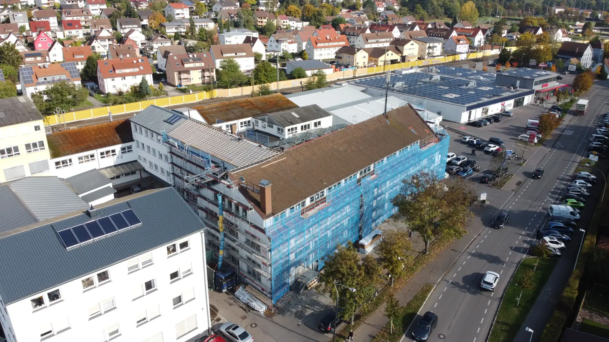 Langgestrecktes Gewerbegebäude mit Gerüst und Schutznetz, dahinter Wohngebiet